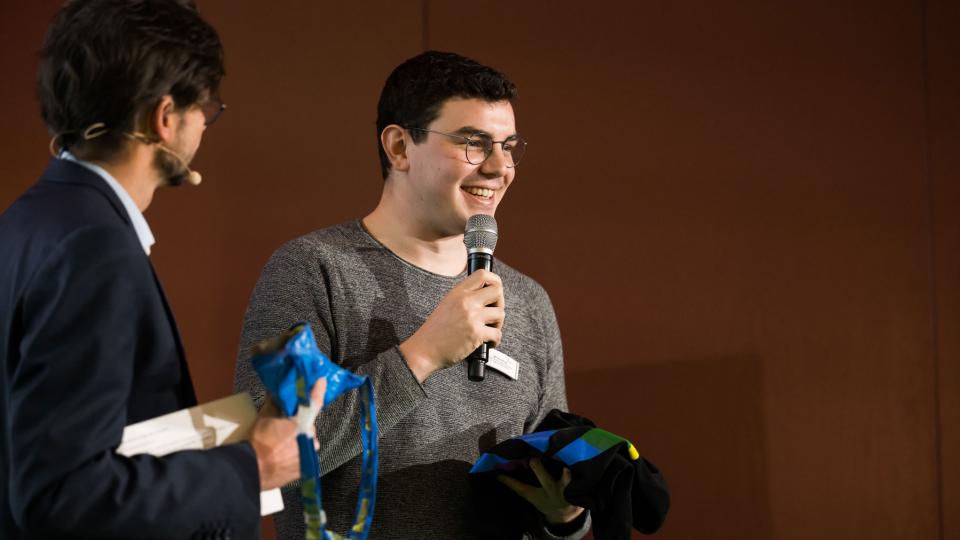 Auf einer Bühne stehen ein Mann mit Headset und einer blauen Tüte in der Hand und ein Mann mit einem T-Shirt und einem Mikrofon in der Hand