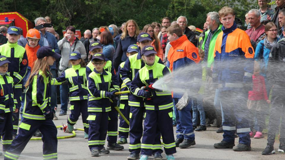 Feuerwehrkinder halten gemeinsam eine Spritze, aus der ein Wasserstrahl schießt.