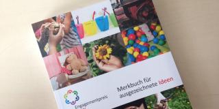 Broschüre mit dem Titel Merkbuch für ausgezeichnete Projekte zum Engagementpreis NRW vor einem beigen Hintergrund. Auf dem Cover sind bunte Fotos mit Gegenständen wie Becher, Teddybären oder alte Telefone.