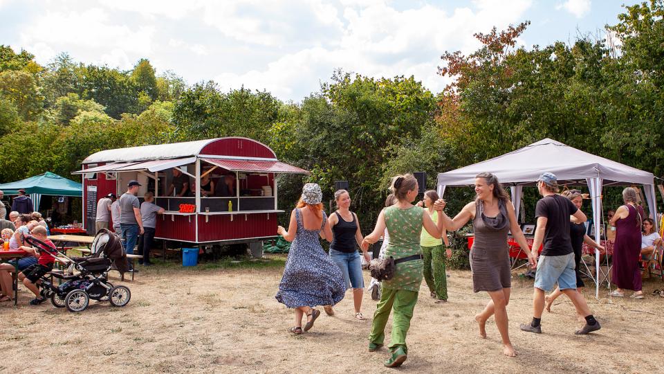 Auf einer Festwiese, die von einem Essensstand und mehreren Zelten sowie Bäumen umringt ist, tanzen Frauen mit lachenden Gesichtern.