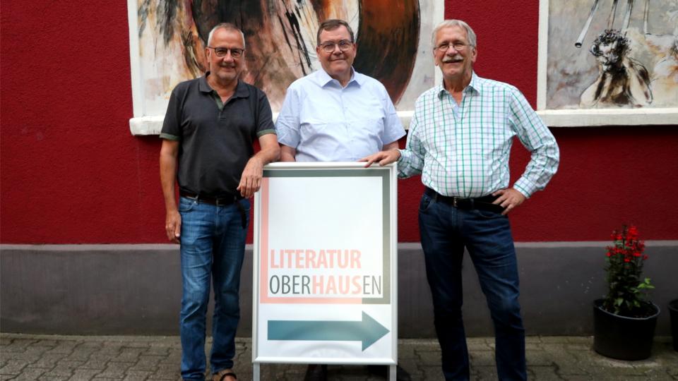 Drei Männer stehen vor einer roten Hauswand an einem Aufsteller, der den Weg zum Literaturhaus Oberhausen weist.