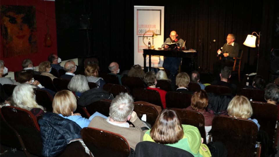 Lesung eines Autors aus einem Buch auf einer Bühne vor einem Publikum in Kinobestuhlung.