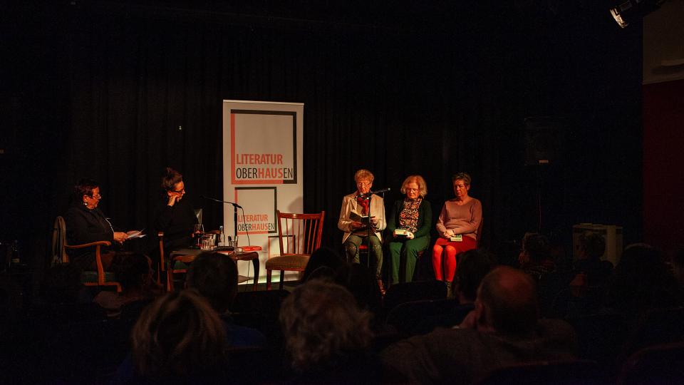 Fünf Frauen sitzen mit Büchern auf einer Theaterbühne. In ihrer Mitte ein Roll-Up des Literaturhauses Oberhausen.