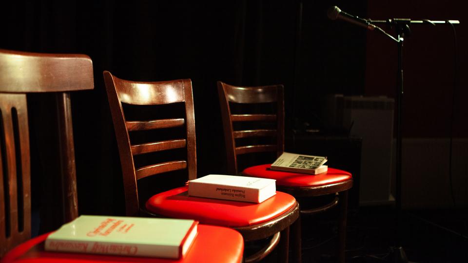 Drei Stühle stehen auf einer dunklen Bühne, es liegt jeweils ein Buch auf der Sitzfläche