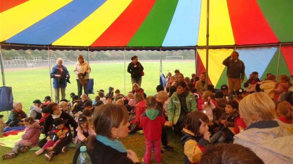 Kinder und Erwachsene unter einem bunten Zirkuszelt auf einer Wiese.