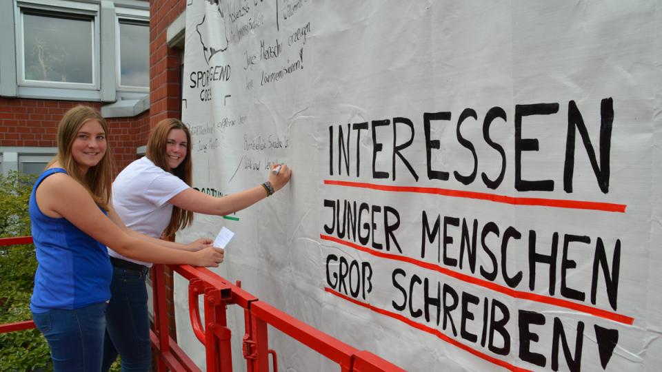 Zwei junge Frauen schreiben auf großem Banner Texte unter dem Slogan "Interessen junger Menschen groß schreiben".