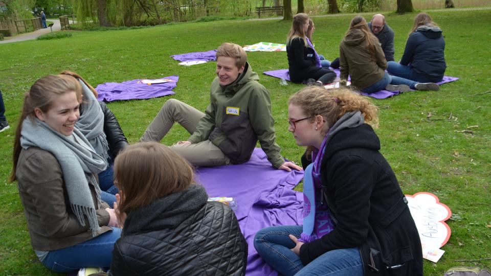 Menschen sitzen auf lila-farbenen Decken in Grünanlage.