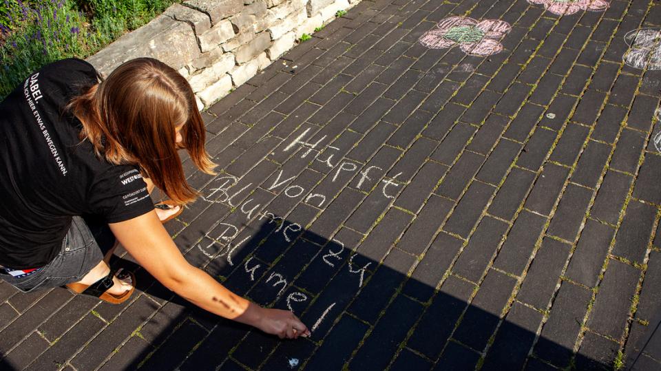 Eine Jugendliche schreibt mit Kreide auf das Pflaster: Hüpfe von Blume zu Blume!