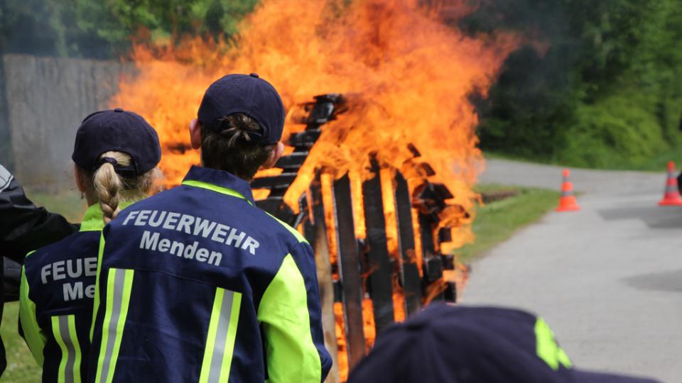 Kinder in Uniformen der Feuerwehr Menden blicken Richtung Holzhütte, die in Flammen steht.