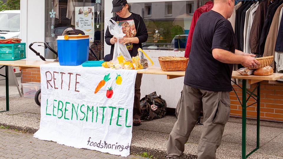 Zwei Personen stehen an Biertischen mit Lebensmitteln drauf, ein selbst erstelltes Banner mit der Aufschrift: »Rette Lebensmittel foodsharing.de« ist am Tisch befestigt