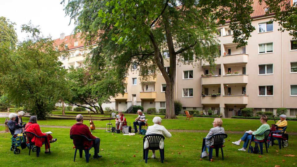 Ältere Menschen sitzen im Halbkreis als Zuschauer bei einem Gartenkonzert auf der Wiese einer Wohnanlage