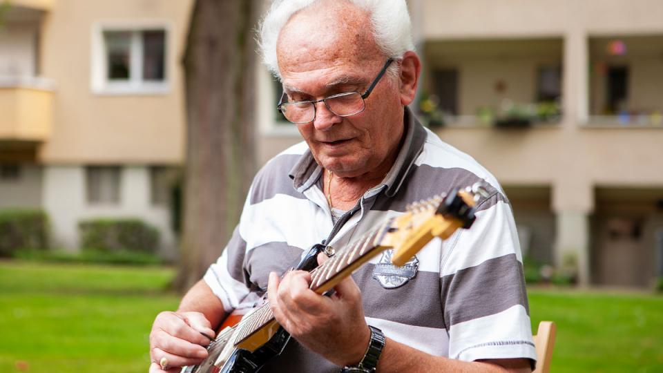 Ein älterer Mann sitzt im Garten vor Wohnanlage und spielt E-Gitarre