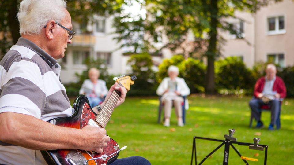 Der Fokus liegt auf einem älteren Mann mit E-Gitarre, weitere Personen sitzen im Garten