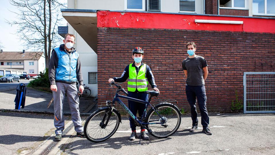 Ein Junge in Warnweste und Mundschutz steht hinter einem Fahrrad, zwei Männer stehen neben ihm
