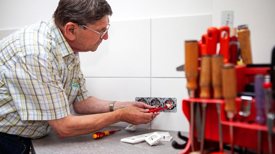 Ein älterer Herr arbeitet mit einem Spannungsprüfer an einer nicht verkleideten Steckdosenleiste.