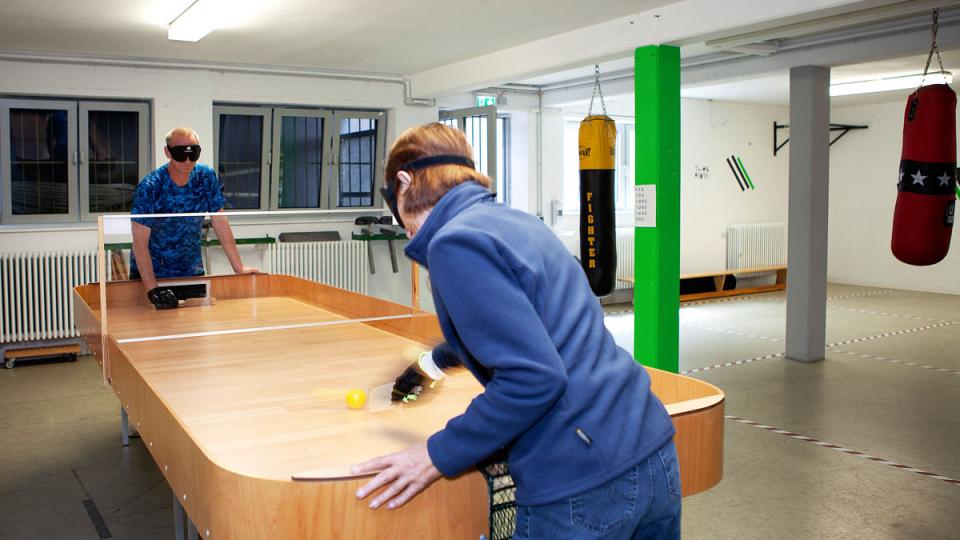 Zwei Menschen mit speziellen Brillen beim Tischball, ein Sportangebot für blinde, sehbehinderte und sehende Menschen