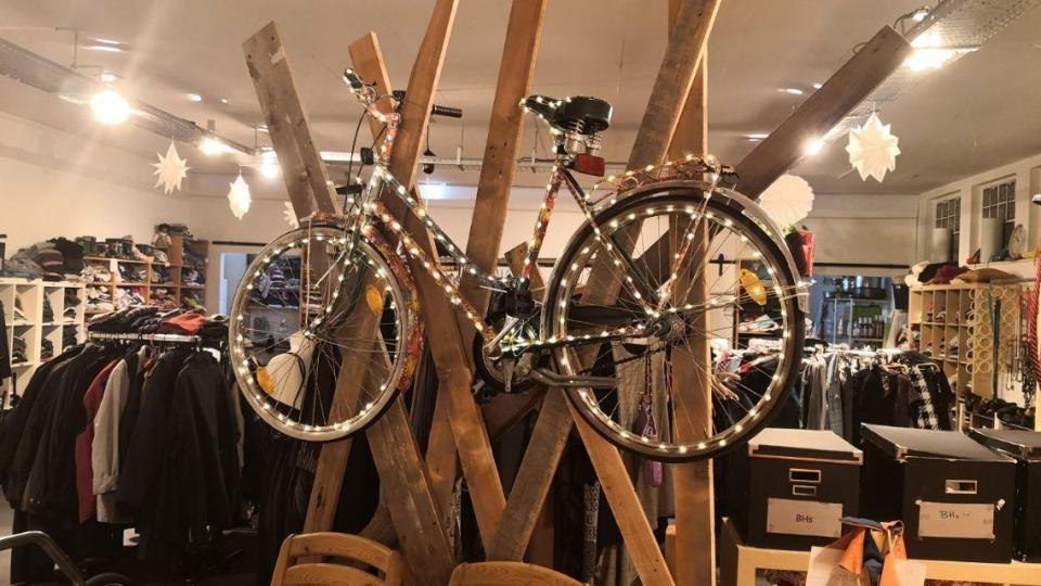 Ein Fahrrad hängt an einer Holzkonstruktion, hinter der ein Kleiderladen zu sehen ist.