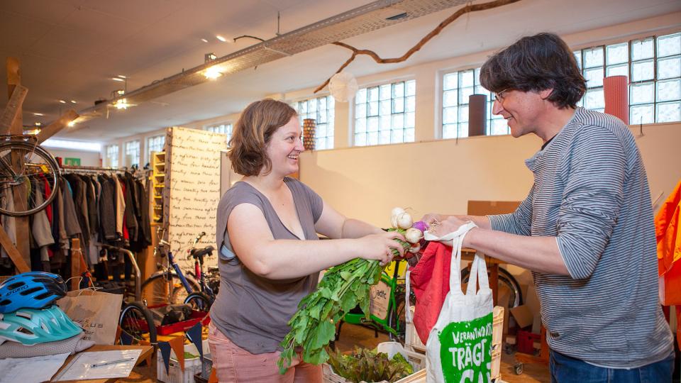 zwei Personen reichen sich Stofftaschen und Gemüse im Fachgeschäft für Stadtwandel