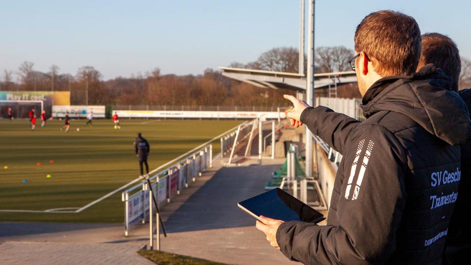 Zwei Trainer zeigen mit einem Tablet in der Hand auf einen Fussballtrainingsplatz