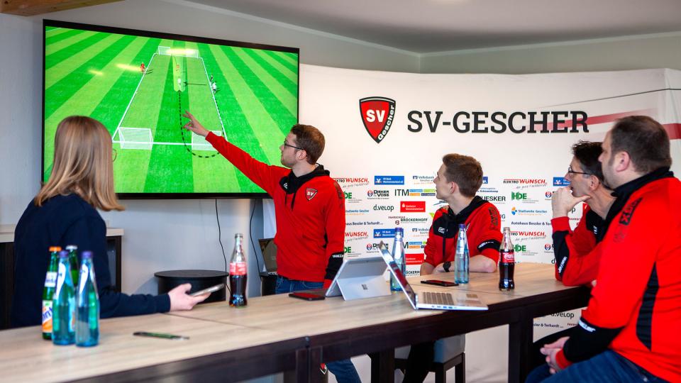 Fünf Personen sitzen am Tisch und schauen auf einen großen Bildschirm, auf dem ein Fussballfeld abgebildet ist