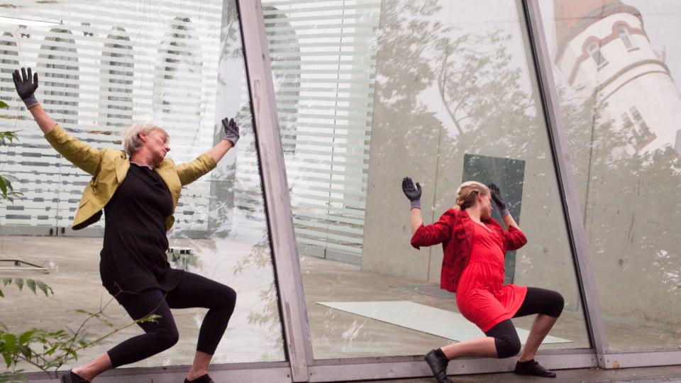 Zwei Frauen lehnen in theatralischer Geste an der Glaswand eines Gebäudes.