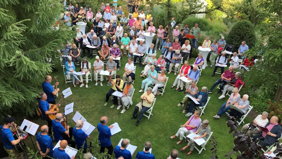 Im Klostergarten spielt eine Gruppe in blauen T-shirts Musik, vor ihnen sitzen Menschen in 2er Gruppen auf weißen Stühlen, im Hintergrund stehen weitere Personen