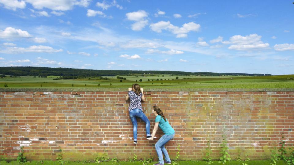 Zwei Mädchen klettern gemeinsam über Mauer, Landschaft Hintergrund mit Wiesen, Wäldern und Himmel