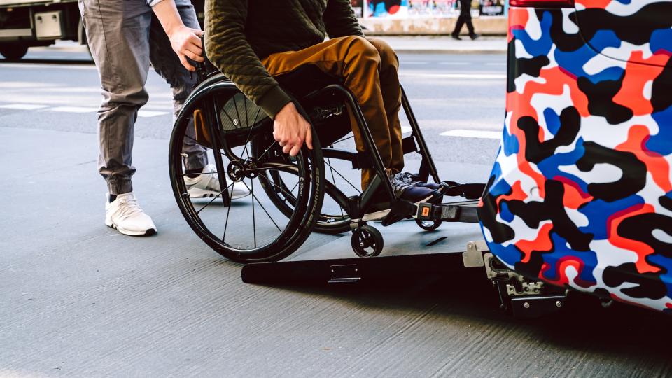 Eine Person auf einem Rollstuhl fährt unter Hilfestellung die Rampe eines Kraftfahrzeugs hoch.