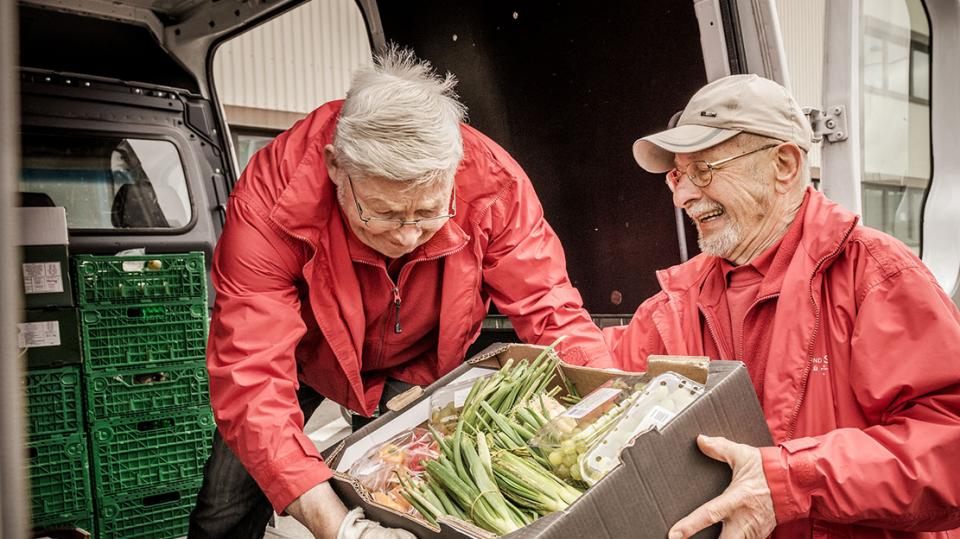 Zwei ältere Menschen heben gemeinsam eine Kiste mit Gemüse in einen Transporter.