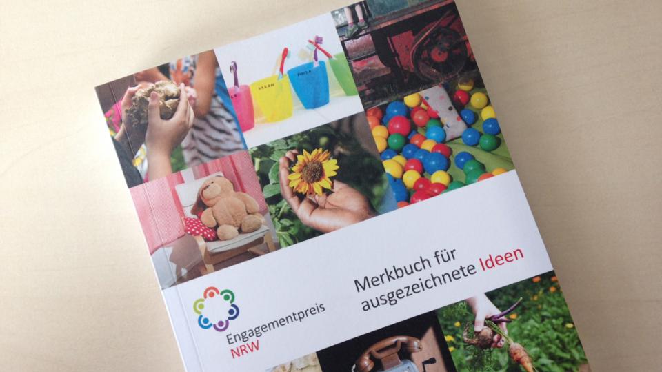 Broschüre mit dem Titel Merkbuch für ausgezeichnete Projekte zum Engagementpreis NRW vor einem beigen Hintergrund. Auf dem Cover sind bunte Fotos mit Gegenständen wie Becher, Teddybären oder alte Telefone.