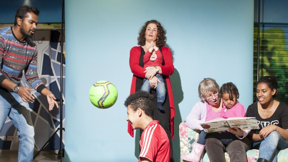 Ein Junge und junger Erwachsener spielen Ball. Eine Frau mit Kind. Eine Seniorin liest zwei Mädchen etwas vor.