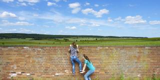 Zwei Mädchen klettern gemeinsam über Mauer, Landschaft Hintergrund mit Wiesen, Wäldern und Himmel