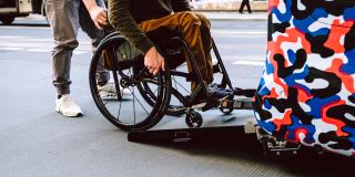 Eine Person auf einem Rollstuhl fährt unter Hilfestellung die Rampe eines Kraftfahrzeugs hoch.