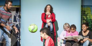 Ein Junge und junger Erwachsener spielen Ball. Eine Frau mit Kind. Eine Seniorin liest zwei Mädchen etwas vor.